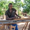 Man in dark t-shirt smiling and standing behind carpentry table 