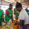 Three people in green clothing handing food and soap to a woman in a white top holding an orange bucket