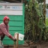 A woman washes her hands in front of a green building with trees in the background