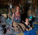 Woman kneels on floor inside a hut with her small baby sitting beside her