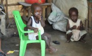 An infant sits in a green plastic chair looking at the camera with her hand over her mouth. Behind her, a second child sits on the floor.
