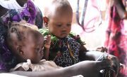 Two infant girls sit in their mother's arms as she tears open a sachet of food for them.