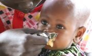 Closeup of a baby sitting on a woman's knee as she feeds her a sachet of food