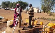 Woman operating hand pump next to man, surrounded by jerry cans 