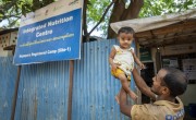 Amir* lifts his one-year-old daughter just outside the Integrated Nutrition Centre in the Nayapara Registered Camp, Teknaf. The vibrant blue signage of the centre marks the familiar locations where families like theirs receive essential nutrition and medical services to address malnutrition and ensure healthy growth among the Rohingya children as well as the pregnant and breastfeeding mothers. (Photo: Saikat Mojumder/Concern Worldwide)