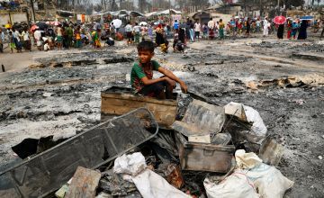 A Rohingya refugee boy sits on a stack of burned material.  