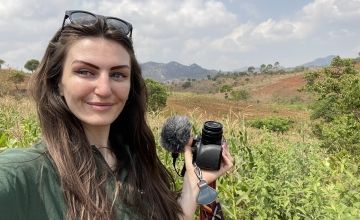 Woman with long brown hair and sunglasses on head holding camera and taking selfie against hills 