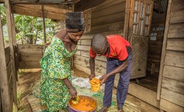 African man and woman scooping food from drum inside wooden cabin