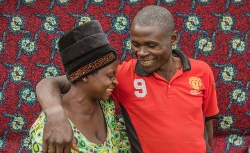 African man in red t-shirt with arm around wife, who is closing eyes and smiling