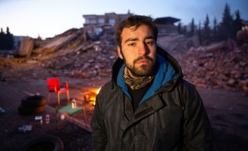 Man with beard in warm jacket stands in front of rubble and fire