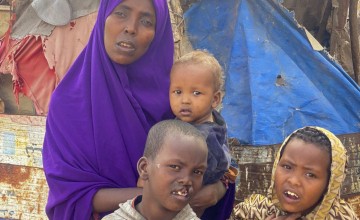 Farhiya, woman from Somaliland, holds infant child with two young boys standing in front of her. She is wearing a purple head scarf