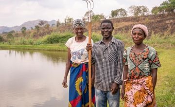 Kosimasi, a farmer, holds a hoe and stands between two female farmers in front of pond in Malawi