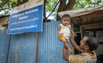 Amir* lifts his one-year-old daughter just outside the Integrated Nutrition Centre in the Nayapara Registered Camp, Teknaf. The vibrant blue signage of the centre marks the familiar locations where families like theirs receive essential nutrition and medical services to address malnutrition and ensure healthy growth among the Rohingya children as well as the pregnant and breastfeeding mothers. (Photo: Saikat Mojumder/Concern Worldwide)