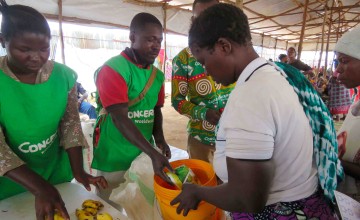 Three people in green clothing handing food and soap to a woman in a white top holding an orange bucket
