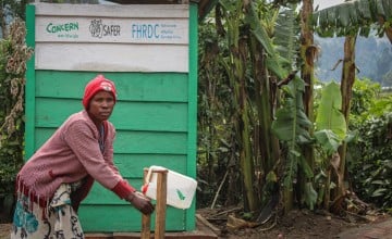 A woman washes her hands in front of a green building with trees in the background