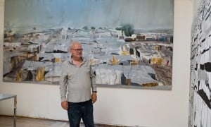 Artist Brian Maguire standing next to two large paintings of Benitu camp in South Sudan