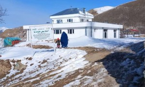 Two people in front of a white two storey building, on snowy rocky ground