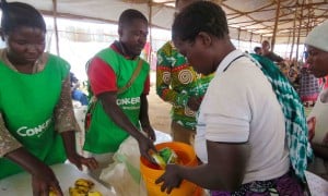 Three people in green clothing handing food and soap to a woman in a white top holding an orange bucket