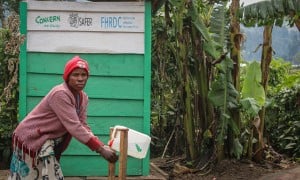 A woman washes her hands in front of a green building with trees in the background