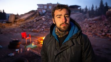 Man with beard in warm jacket stands in front of rubble and fire