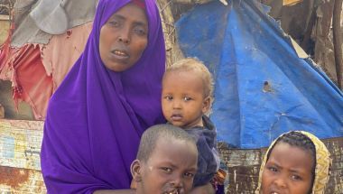 Farhiya, woman from Somaliland, holds infant child with two young boys standing in front of her. She is wearing a purple head scarf