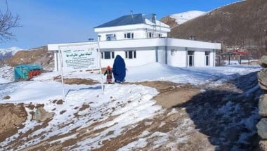 Two people in front of a white two storey building, on snowy rocky ground