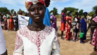 Ajok Deng, a woman in South Sudan wears a white dress, holding some cash. Behind her is a queue of people
