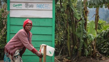 A woman washes her hands in front of a green building with trees in the background