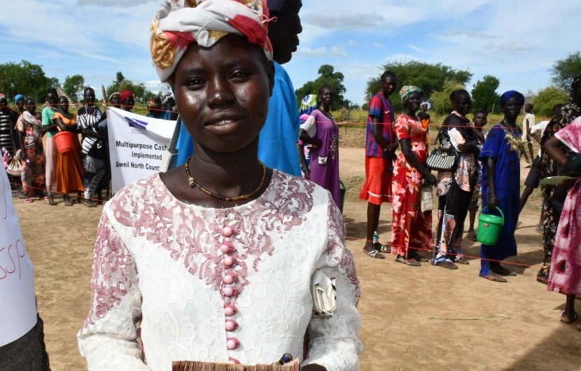 Ajok Deng, a woman in South Sudan wears a white dress, holding some cash. Behind her is a queue of people
