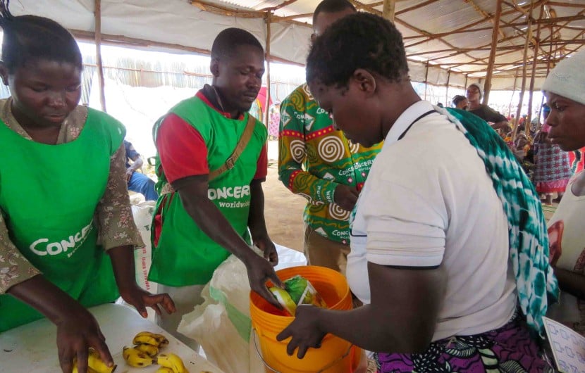 Three people in green clothing handing food and soap to a woman in a white top holding an orange bucket