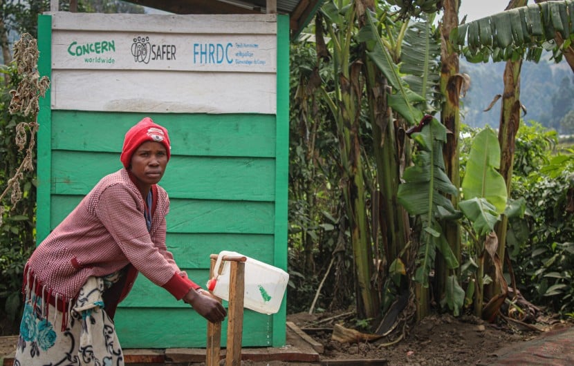 A woman washes her hands in front of a green building with trees in the background