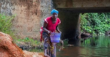 Woman collects water in bright plastic bucket.