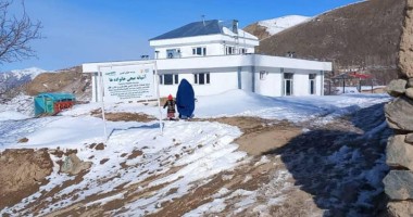 Two people in front of a white two storey building, on snowy rocky ground