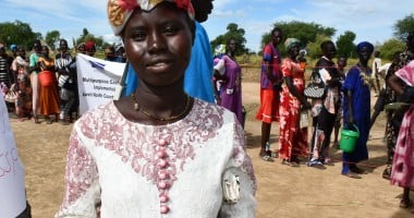 Ajok Deng, a woman in South Sudan wears a white dress, holding some cash. Behind her is a queue of people