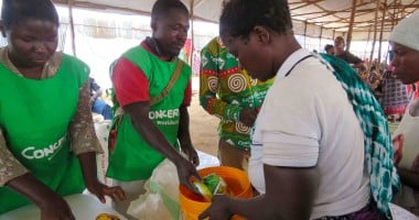 Three people in green clothing handing food and soap to a woman in a white top holding an orange bucket