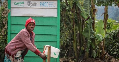 A woman washes her hands in front of a green building with trees in the background