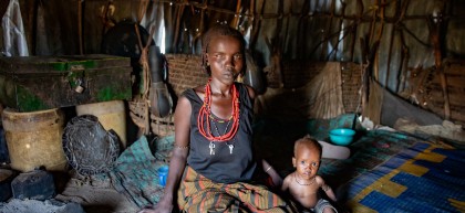 Child sits on floor of hut beside his mother, both look at the camera