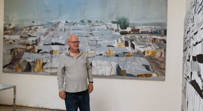 Artist Brian Maguire standing next to two large paintings of Benitu camp in South Sudan