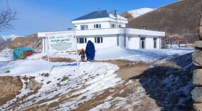 Two people in front of a white two storey building, on snowy rocky ground