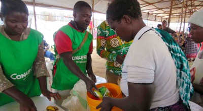 Three people in green clothing handing food and soap to a woman in a white top holding an orange bucket