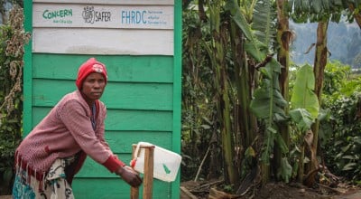 A woman washes her hands in front of a green building with trees in the background